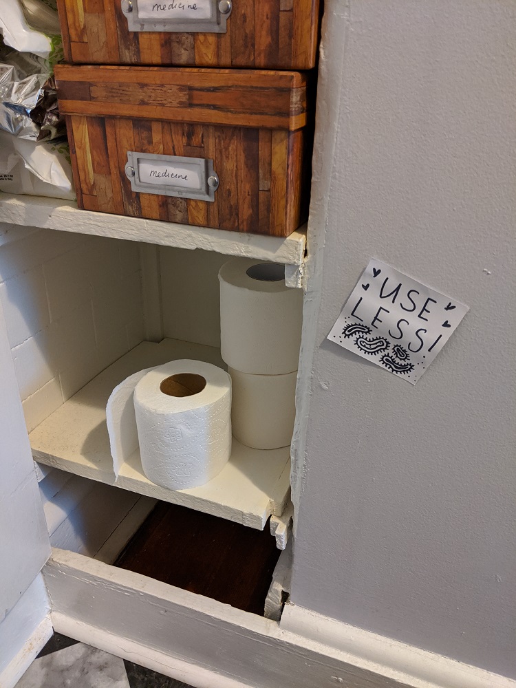 Three rolls of toilet paper on a shelf. Handwritten sign that says 'use less!'