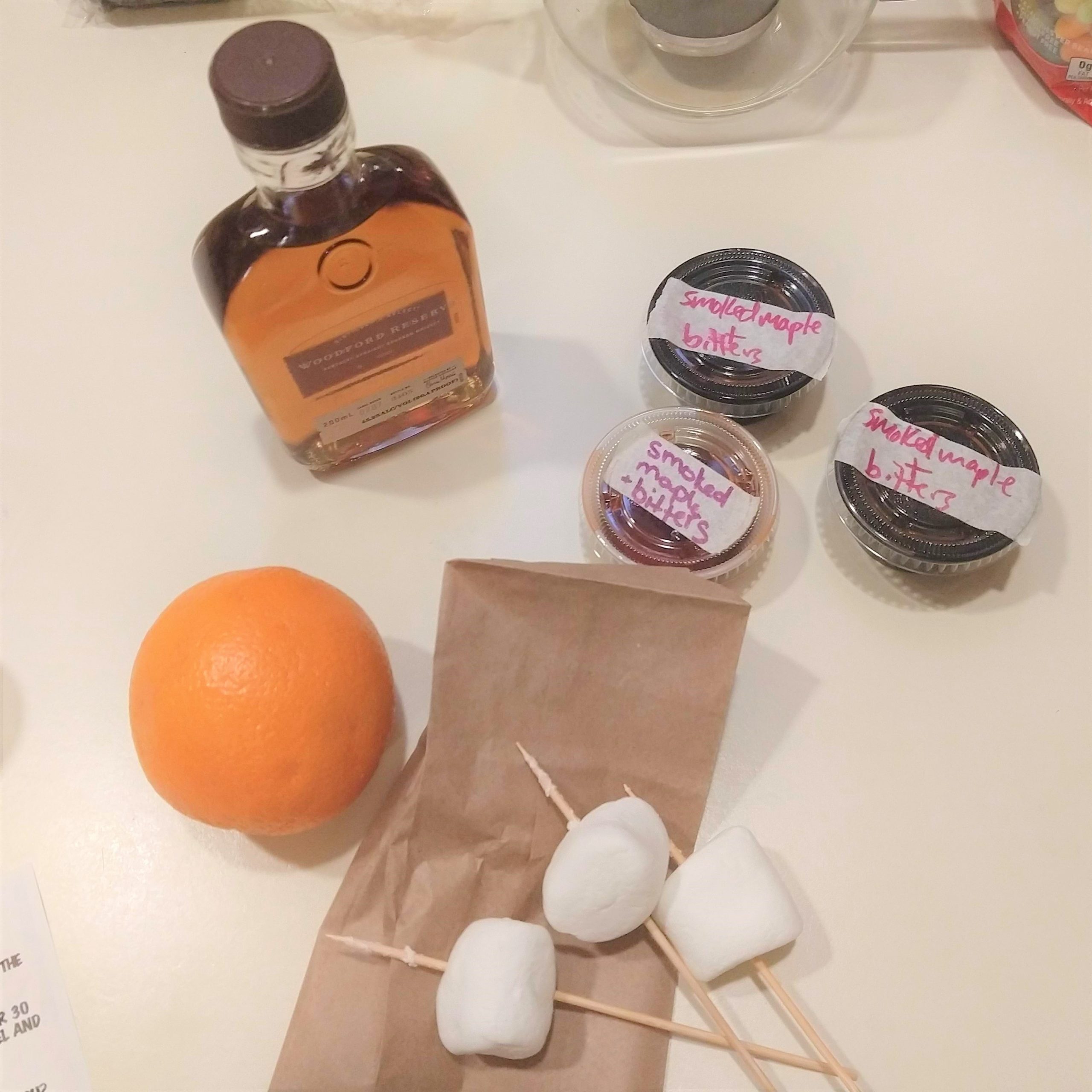 Small bottle of alcohol, three small plastic containers, an orange, and three marshmallows on sticks.